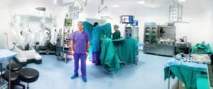 Prof. Dr. Uğur Boylu ameliyathanede Da Vinci robotunun önünde duruyor. Arka planda ameliyat masasında bir doktor ve hemşire ameliyat hazırlığı yapıyor.